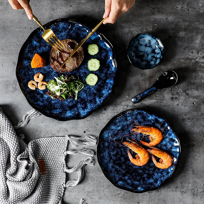 Two different sized, blue textured ceramic plates on a dark table, one with a meal being eaten with gold utensils and the other with three shrimp. A matching small bowl and spoon are also on the table.