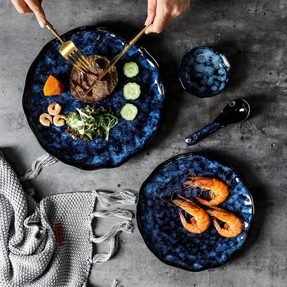 Two different sized, blue textured ceramic plates on a dark table, one with a meal being eaten with gold utensils and the other with three shrimp. A matching small bowl and spoon are also on the table.