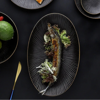A long, dark oval plate with a ridged texture and gold rim holds a grilled fish garnished with lettuce and a lemon slice, placed on a dark surface next to other plates and cutlery.