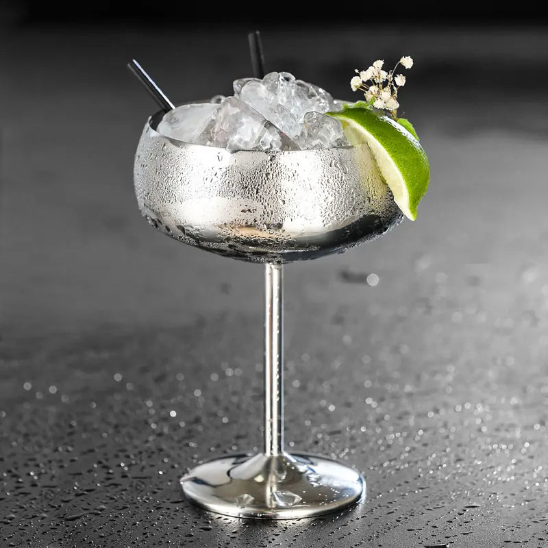 A chilled silver cocktail glass with a lime wedge, ice, and two straws.