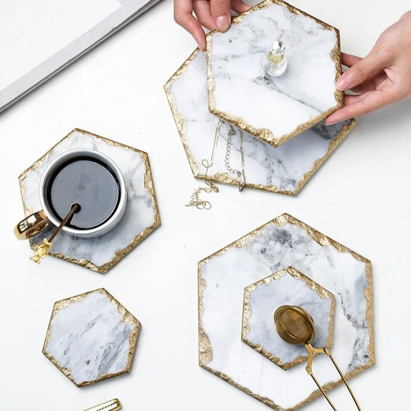 Hands placing a hexagon marble coaster on a table with other coasters, a cup of coffee, and a tea infuser.