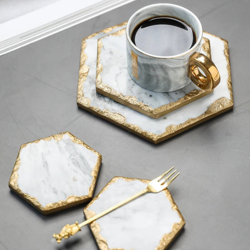A white marble cup with a gold handle filled with coffee, resting on a marble coaster, next to two other marble coasters and a gold fork.
