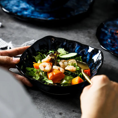 A person's hands holding a dark blue textured ceramic bowl filled with a salad of greens, shrimp, sweet potato, and cucumbers, with a gold utensil.