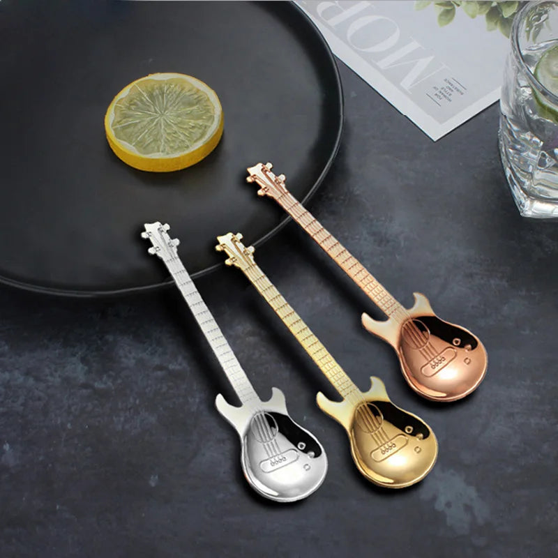 Three guitar-shaped spoons in silver, gold, and rose gold on a dark surface with a lemon wedge.