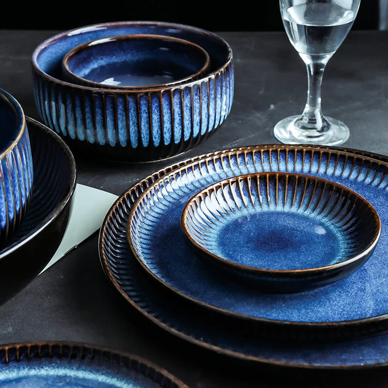 A table is set with multiple pieces of shiny, blue ceramic dinnerware, including nested bowls and plates.