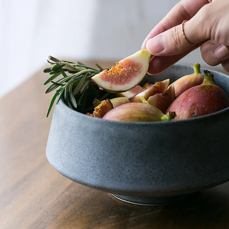 A hand holding a sliced fig above a dark ceramic bowl filled with fresh figs and a sprig of rosemary on a wooden table. The scene highlights natural textures and earthy tones.