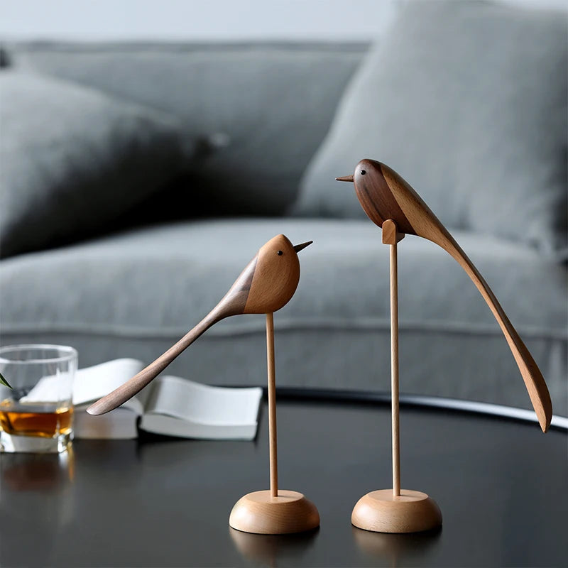 Two decorative wooden bird figurines with long tails on wooden stands, set on a dark coffee table with a glass of amber liquid, in front of a gray sofa.