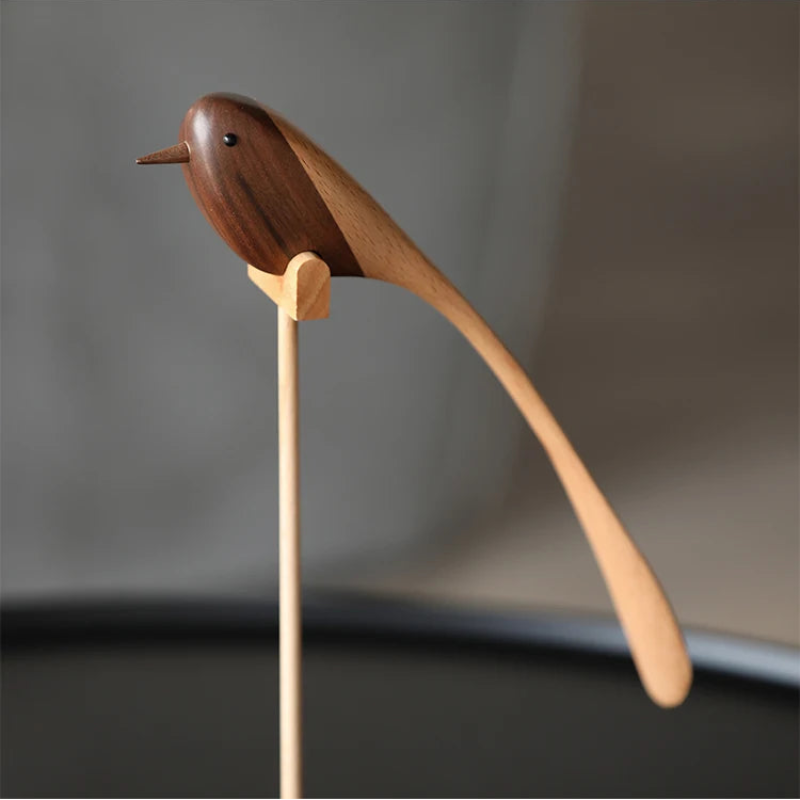 A single decorative wooden bird figurine with a long, sweeping tail, made from contrasting dark and light wood, perched on a thin wooden stand.