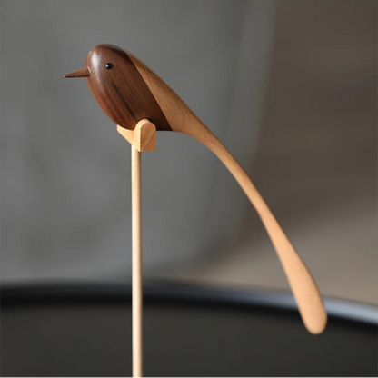 A single decorative wooden bird figurine with a long, sweeping tail, made from contrasting dark and light wood, perched on a thin wooden stand.