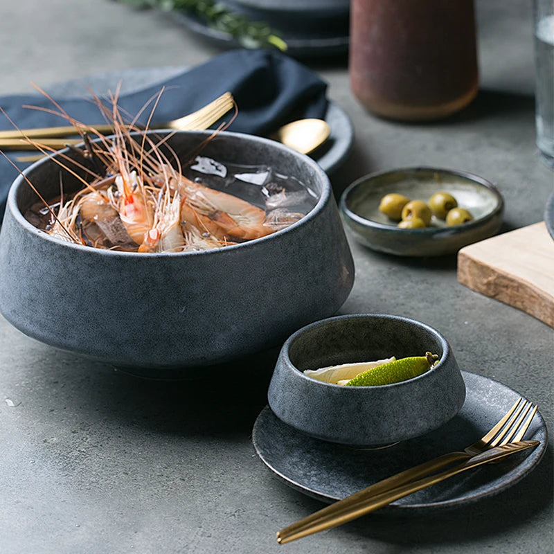 A dark ceramic bowl filled with fresh shrimp on ice, accompanied by a smaller bowl with lime wedges and butter. The setting includes gold utensils, a navy napkin, and side dishes with olives, all arranged on a textured gray surface.