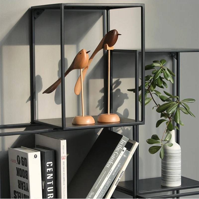 Two decorative wooden bird figurines with long tails on wooden stands are displayed on an open black metal shelving unit above a stack of books and a potted plant in a striped vase.