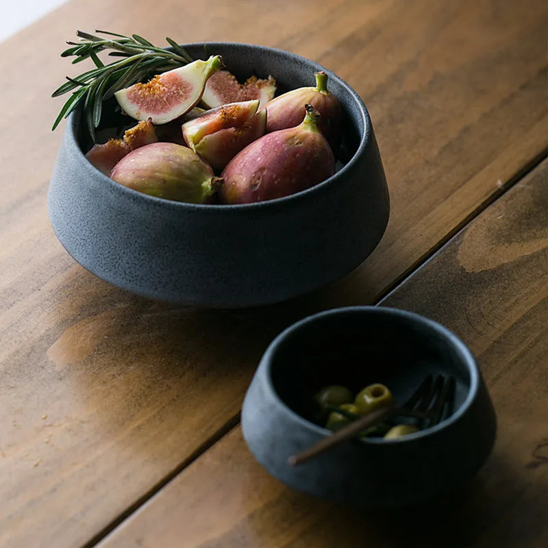 A dark ceramic bowl filled with fresh figs and a sprig of rosemary on a wooden table. A smaller bowl with green olives and a fork sits nearby, creating a rustic and elegant dining scene.