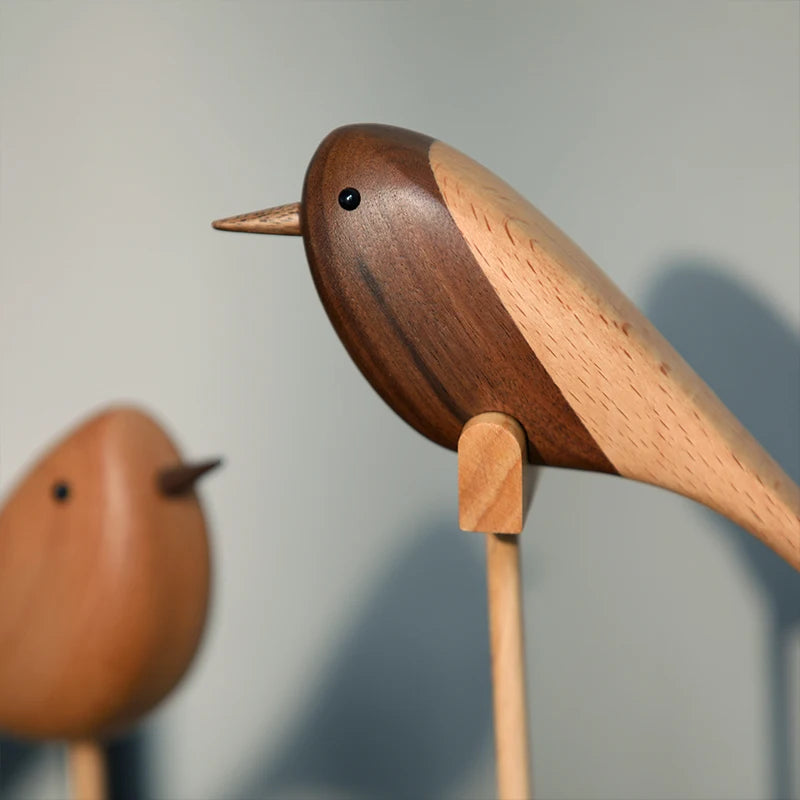 Close-up of a decorative wooden bird figurine made from two contrasting shades of wood, perched on a wooden dowel; a second, lighter bird is visible in the soft background.