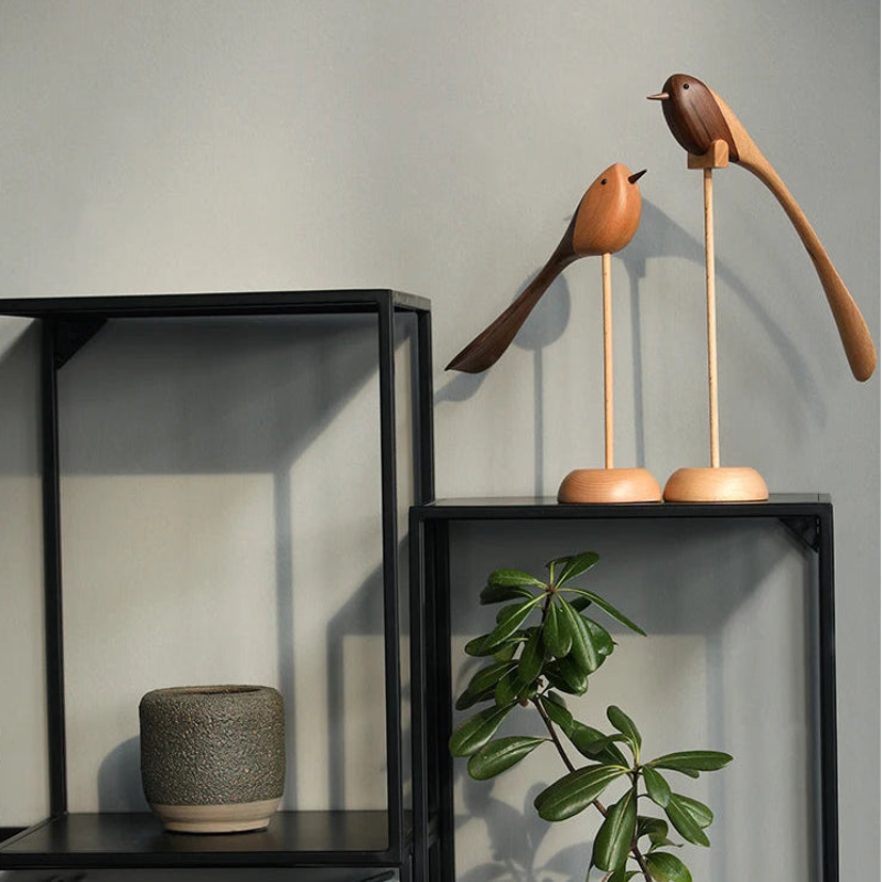 Two decorative wooden bird figurines with long tails on wooden stands are displayed on a black metal shelf unit with a small plant and textured pot.