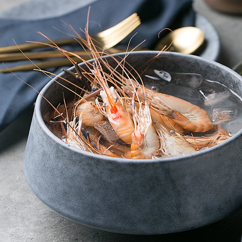 A dark ceramic bowl filled with fresh shrimp on ice, garnished with peppercorns. Gold utensils and a navy napkin are in the background, adding to the elegant dining setup.