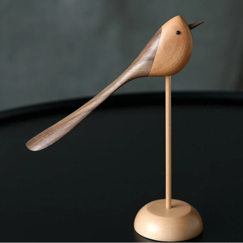 A single decorative wooden bird figurine with a long, sweeping tail, made from contrasting light and dark wood, standing on a thin dowel and round base on a dark table.