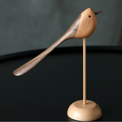 A single decorative wooden bird figurine with a long, sweeping tail, made from contrasting light and dark wood, standing on a thin dowel and round base on a dark table.