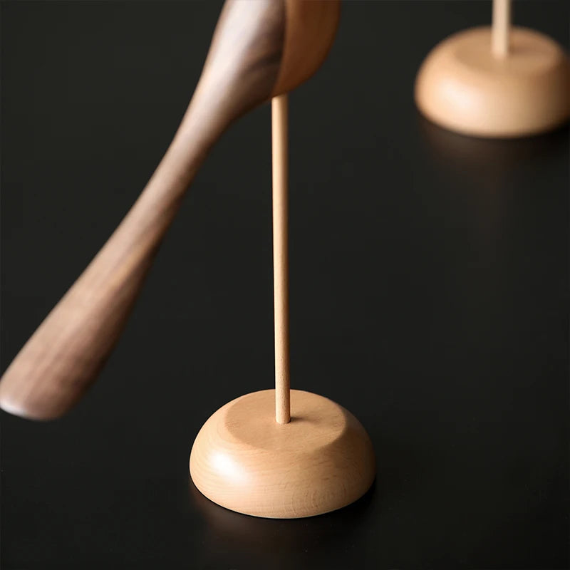 Close-up of the light wood round base and thin dowel holding up a dark wooden bird figurine with a long tail on a dark surface.