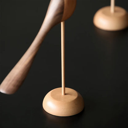 Close-up of the light wood round base and thin dowel holding up a dark wooden bird figurine with a long tail on a dark surface.