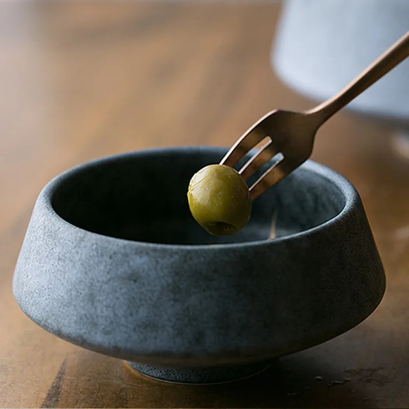 A dark ceramic bowl on a wooden table with a single green olive held by a gold-toned fork. The minimalist setting emphasizes texture and contrast.