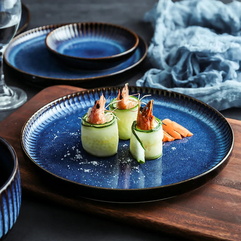 A blue ceramic dinner plate with fluted edges holds an appetizer of shrimp wrapped in cucumber slices.