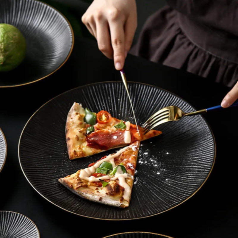 A person's hands use gold cutlery to cut a slice of pizza on a black, ridged plate with a gold rim, set on a dark table.