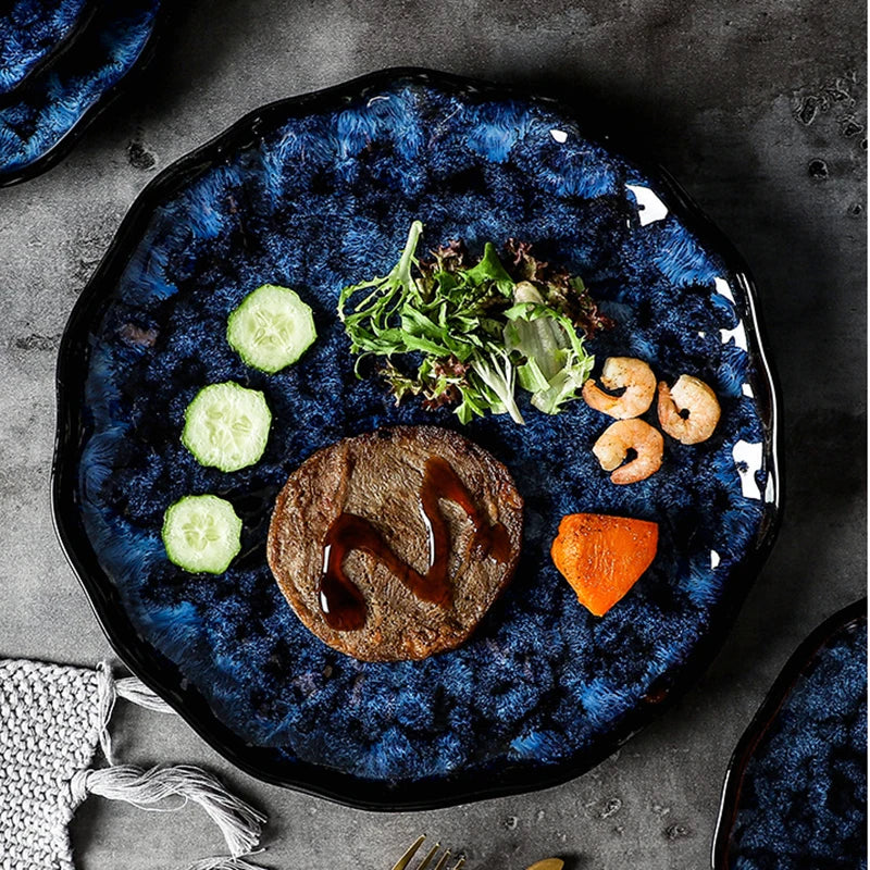A blue ceramic plate with a textured, wavy edge, holding a meal of a burger patty, salad, cucumber slices, shrimp, and a piece of sweet potato.