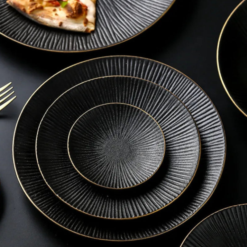 Three black ceramic plates with a ridged texture and gold rims are stacked on a dark surface.