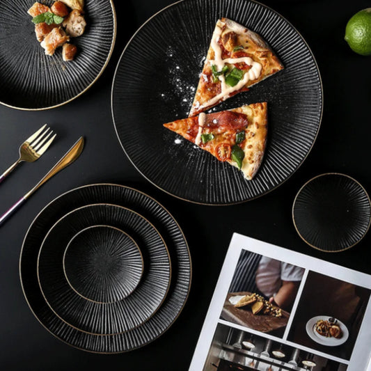 Black dishes with gold rims hold pizza and other food items, arranged on a dark surface with cutlery and a magazine.