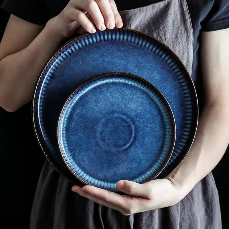 A person holds a pair of nested, deep blue ceramic plates with fluted edges.