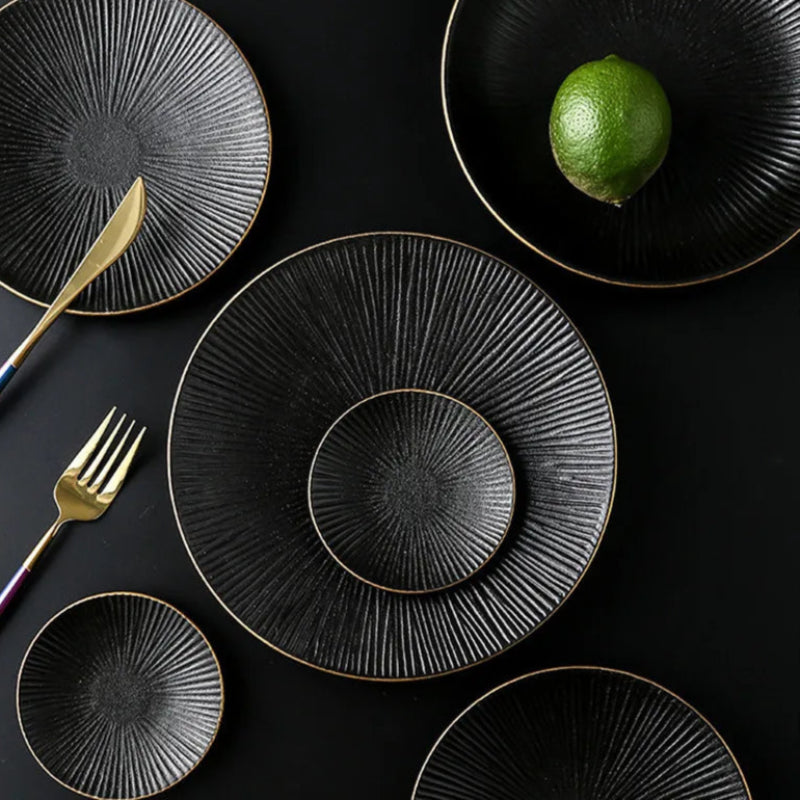 A top-down view shows various black ceramic plates with gold rims and a ridged pattern, arranged on a dark surface with gold cutlery and a single lime.