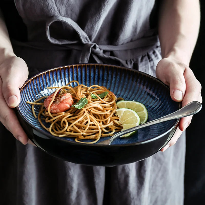A person holds a deep, blue ceramic bowl filled with pasta, a single shrimp, and lime wedges.