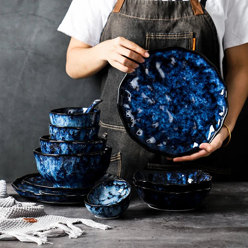 A person in an apron holding a large, blue and white, textured ceramic plate. A matching set of stacked bowls and other dishes is on the table next to them.