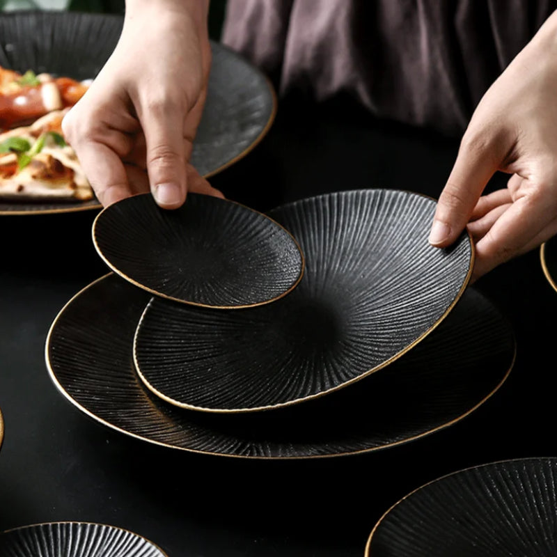 A person's hands arrange three black ceramic plates with ridged textures and gold rims, stacking them on a dark surface.