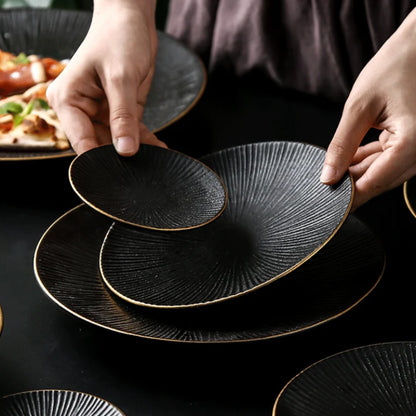 A person's hands arrange three black ceramic plates with ridged textures and gold rims, stacking them on a dark surface.