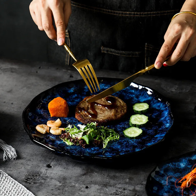 A close-up of a person using a gold fork and knife to eat a meal from a dark blue textured ceramic plate. The meal includes a patty, salad, shrimp, sweet potato, and cucumbers.