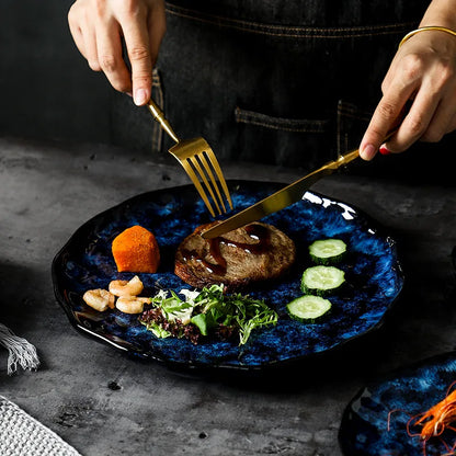A close-up of a person using a gold fork and knife to eat a meal from a dark blue textured ceramic plate. The meal includes a patty, salad, shrimp, sweet potato, and cucumbers.