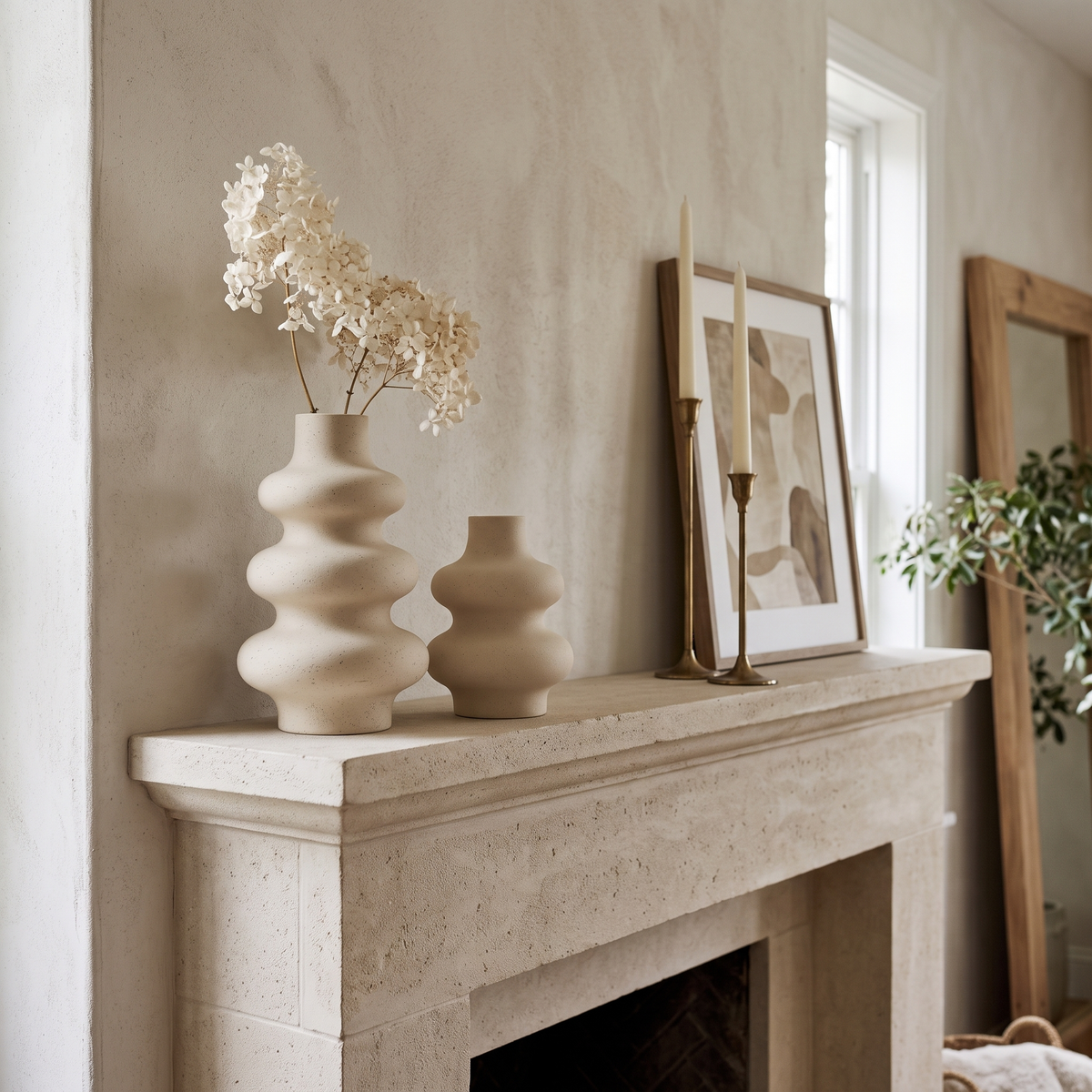 Cream rippled ceramic vase holding dried white flowers beside a smaller vase on a stone fireplace mantel with a framed print