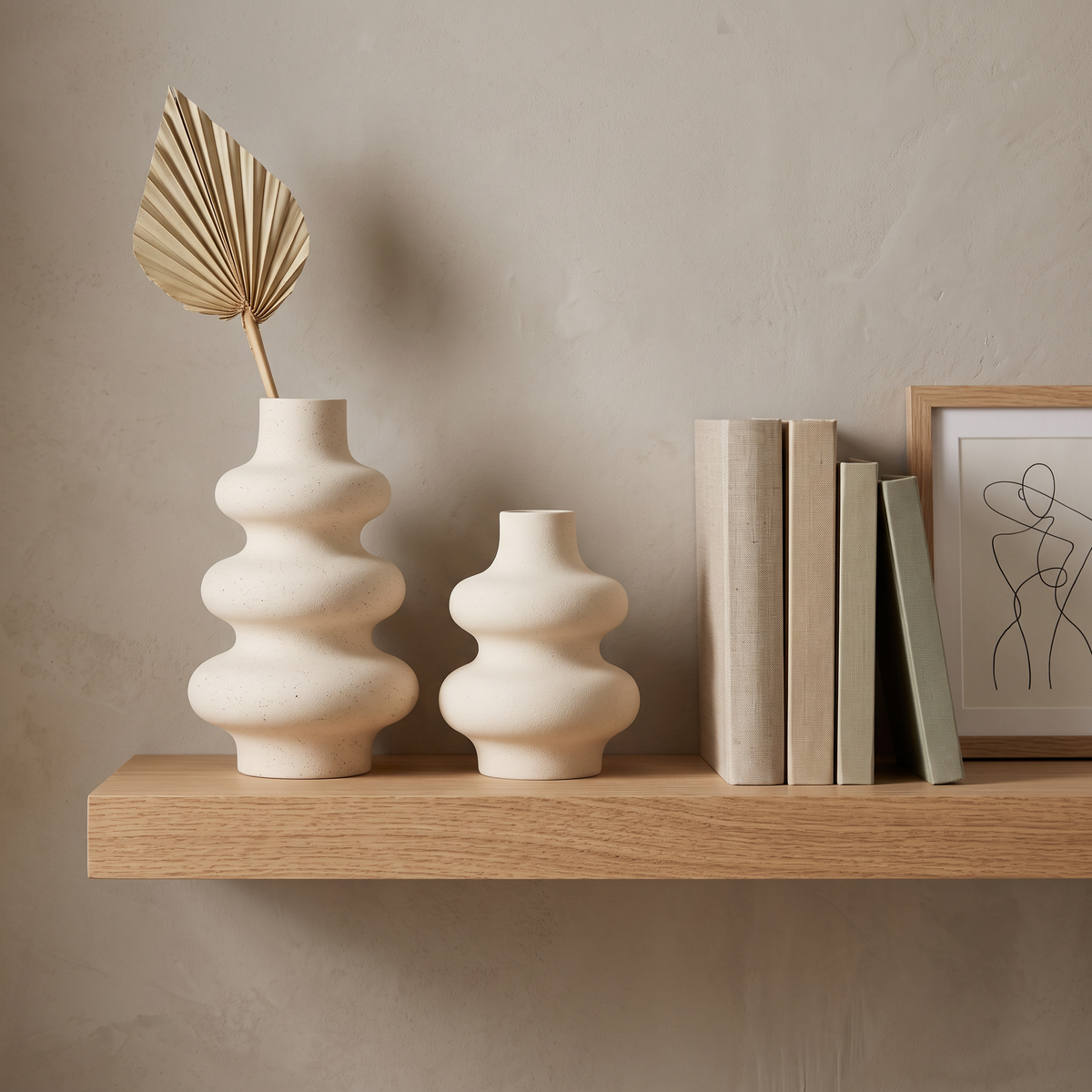 Two cream rippled ceramic vases of different sizes on a wood wall shelf, one holding a dried fan palm leaf, beside a row of books and a framed line drawing