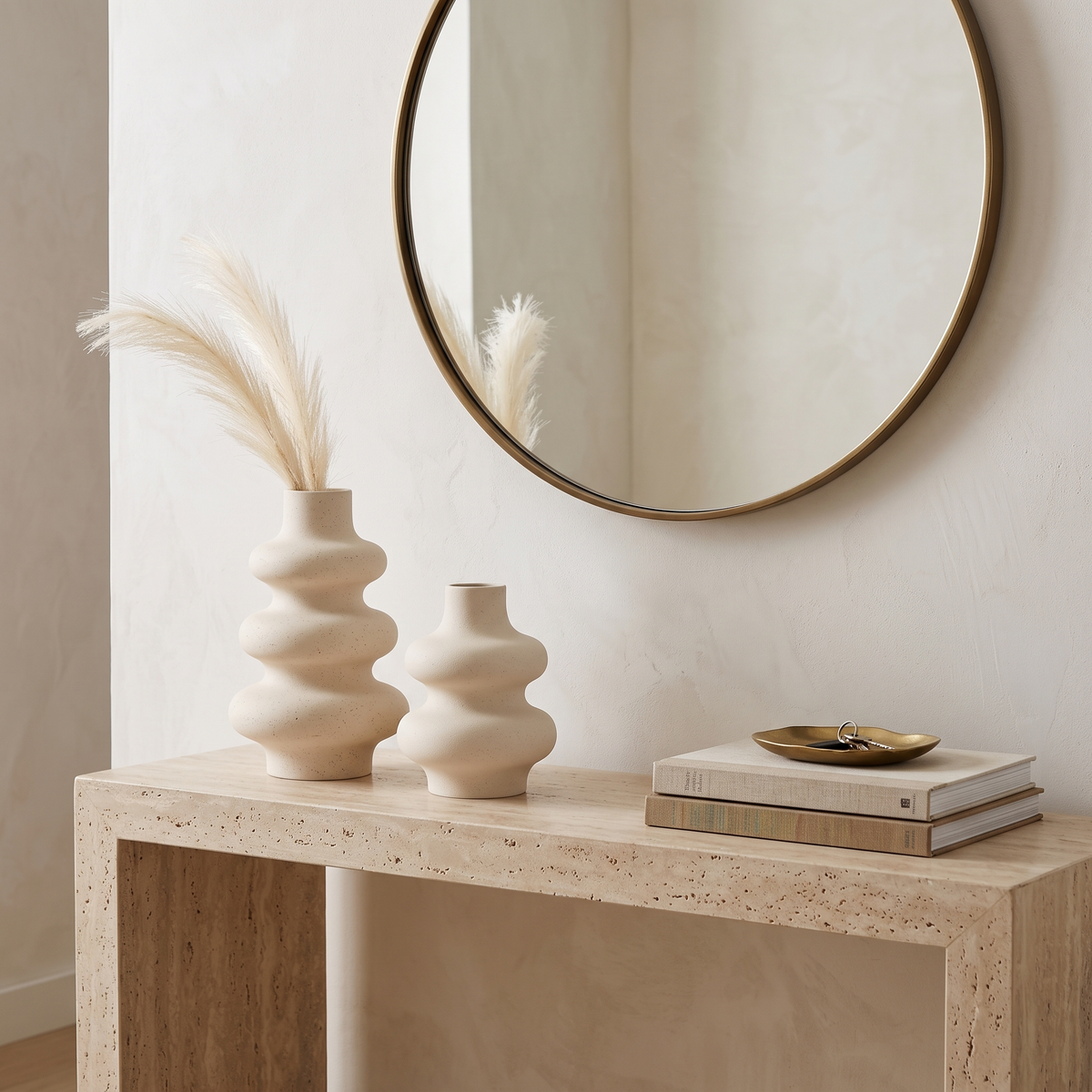 Two cream rippled ceramic vases of different sizes on a light wood console table holding dried bunny tail grass, beside a stack of books and a small dish, with a round mirror above