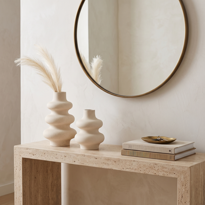 Two cream rippled ceramic vases of different sizes on a light wood console table holding dried bunny tail grass, beside a stack of books and a small dish, with a round mirror above