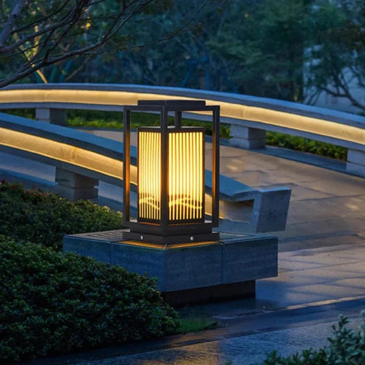A modern, dark-framed outdoor lantern with vertical slats emits a warm glow as it sits atop a square stone pedestal in a garden at dusk or dawn. A curved, illuminated walkway or bridge is visible in the blurred background, along with lush greenery.