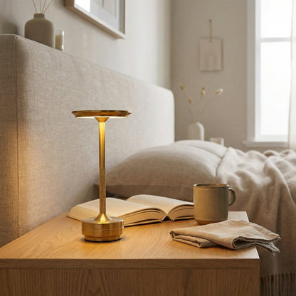 Gold lamp on a wooden nightstand with an open book and mug in a cozy bedroom setting.