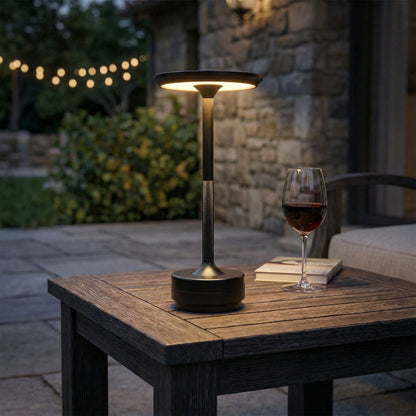 Outdoor setting with a lamp and glass of wine on a wooden table, surrounded by garden lights and stone wall.