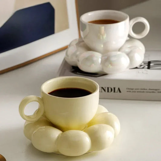 Two ceramic mugs with cloud-shaped saucers, one white and one yellow, with coffee in them. The word "INDIA" is visible in the background.