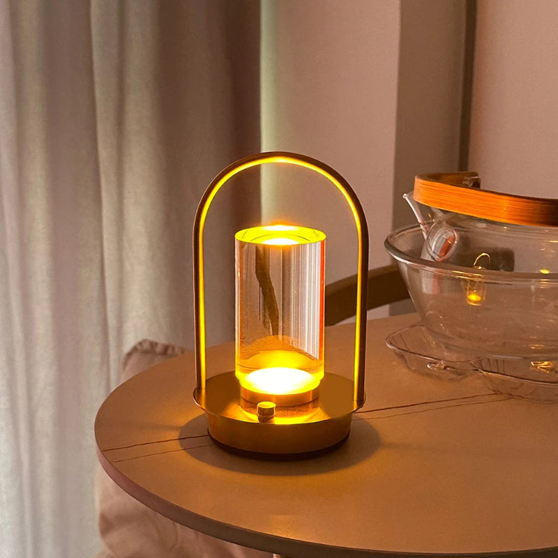 A lit, modern, portable table lamp with a gold metal frame and a glass shade, sitting on a round side table.