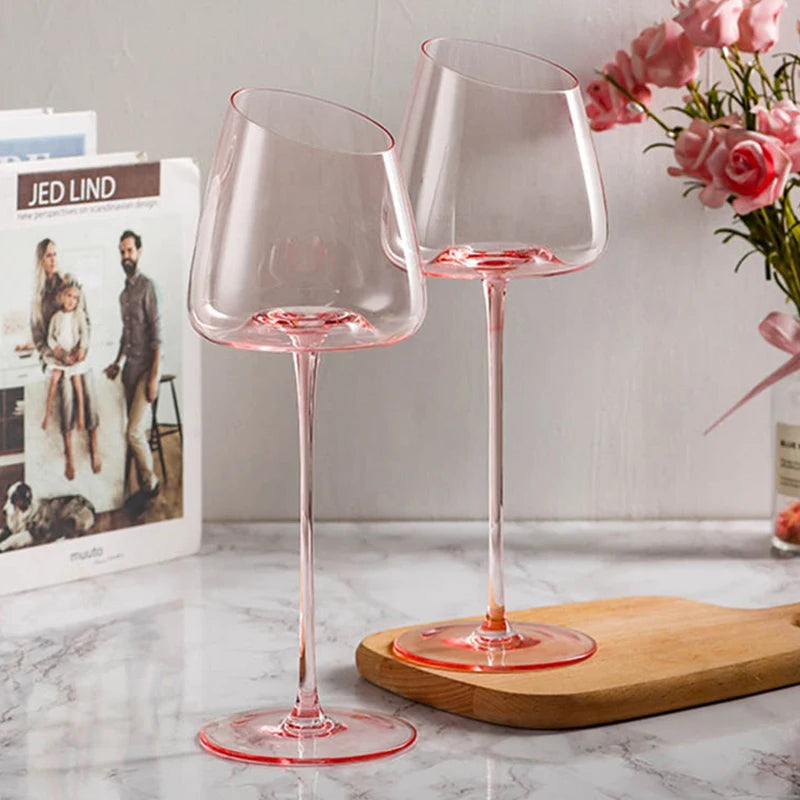 Two pink-stemmed wine glasses with an angled rim on a marble surface, next to a book and a flower vase.