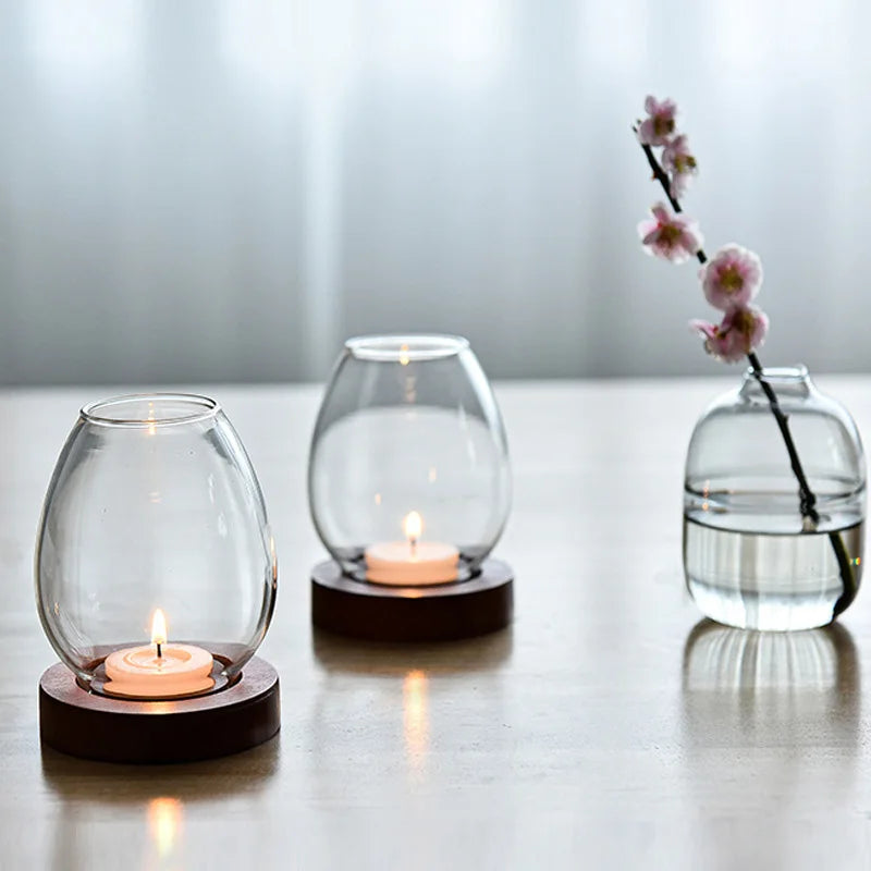 Two oval glass candle holders with wooden bases, each holding a lit tea light candle, placed on a table. A small glass vase with water and a blooming flower branch is positioned nearby, creating a cozy and elegant ambiance.