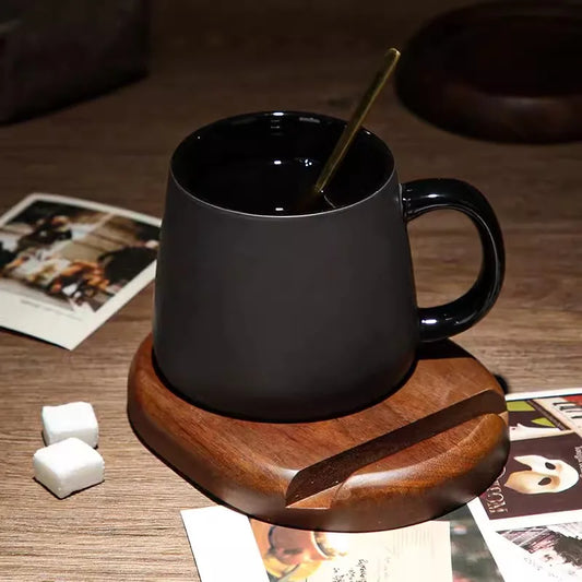 A matte black ceramic mug with a black handle and interior, holding a gold spoon, sits on a dark wooden coaster. Two white sugar cubes and several scattered polaroid photos are nearby.