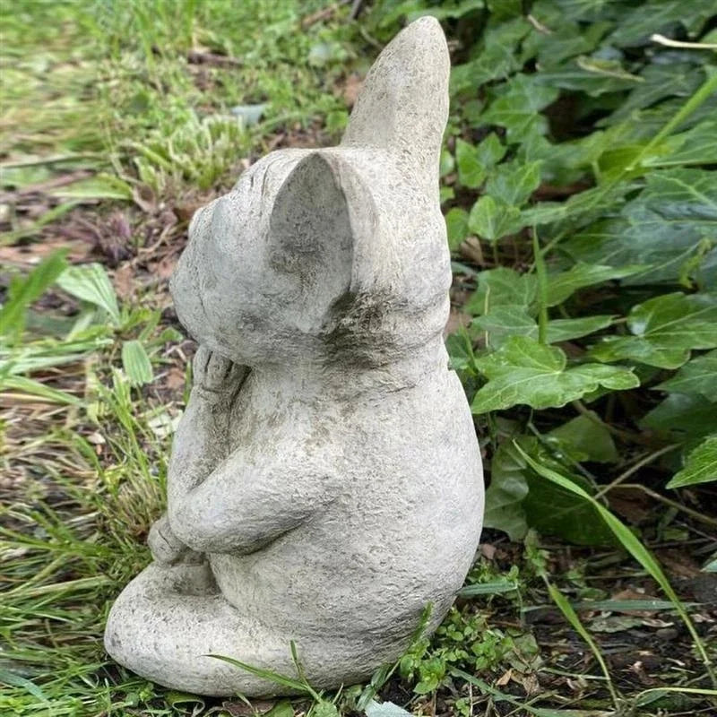 A gray stone statue of a French Bulldog sits cross-legged in a garden, resting its head on its paw in a thoughtful pose, surrounded by green grass and ivy.